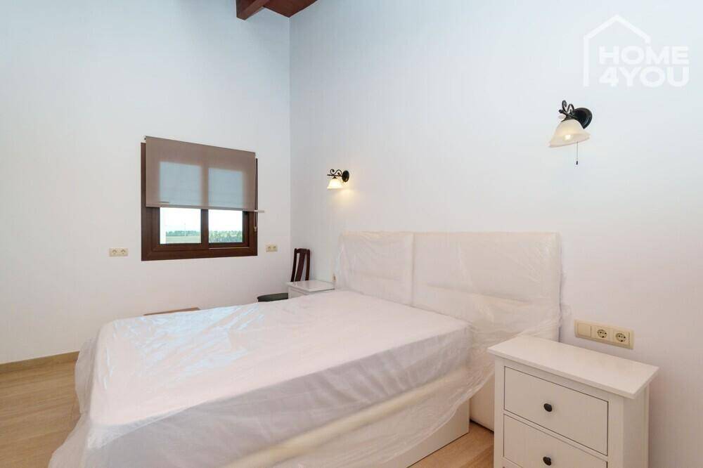 Schlafzimmer mit weißem Bett, Nachttisch und Fenster, das viel Tageslicht hereinlässt.