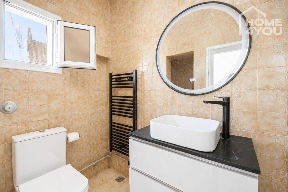 Modernisiertes Badezimmer mit beigen Fliesen, rundem Spiegel und schwarzer Armatur.