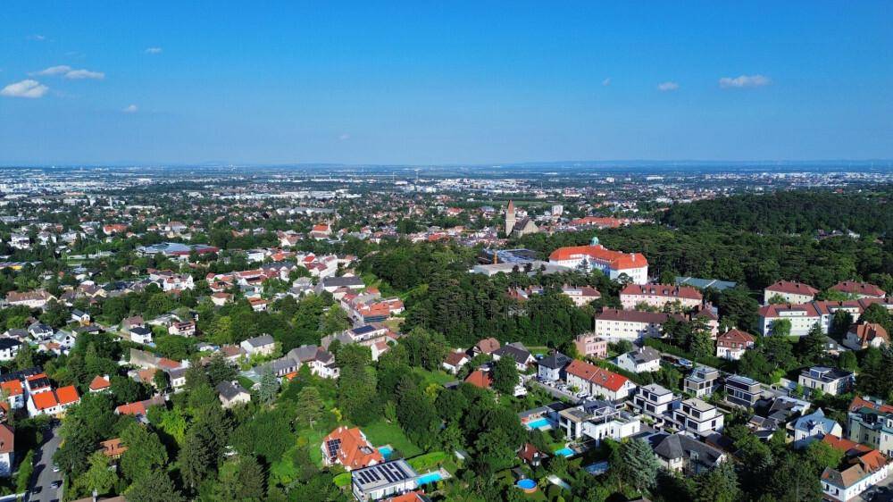 Weitläufige Luftaufnahme einer grünen Wohngegend mit vielen Häusern und Bäumen unter blauem Himmel.