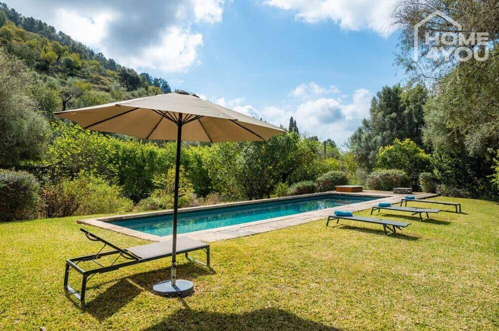 Langer Swimmingpool im grünen Garten mit Sonnenliegen und Sonnenschirm unter blauem Himmel.