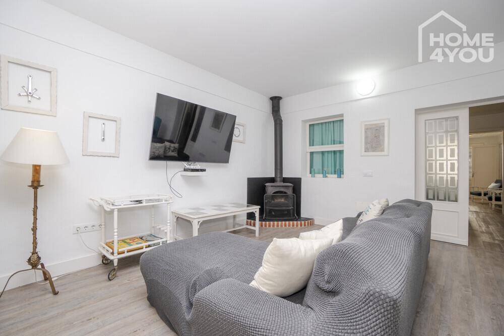 Gemütliches Wohnzimmer mit grauem Sofa, Wandfernseher und einem traditionellen Holzofen.
