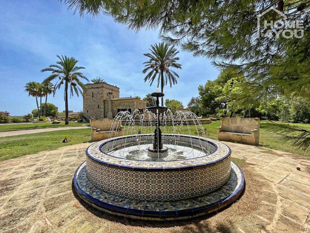 Dekorativer Brunnen mit Mosaikfliesen im weitläufigen Garten, mit dem traditionellen Haus im Hintergrund.