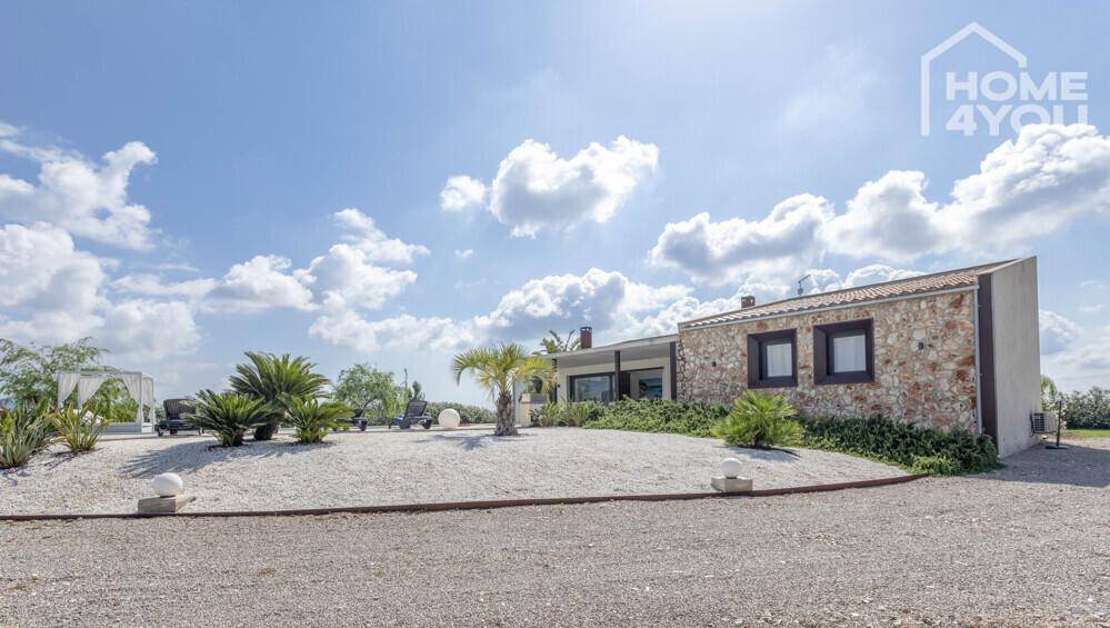 Hausfassade mit Natursteinverkleidung und einem Kiesweg, umgeben von mediterranen Pflanzen.