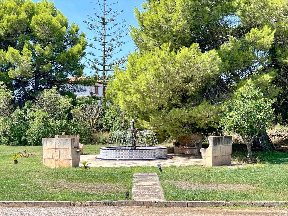 Gepflegter Gartenbereich mit einem dekorativen Brunnen und Steinbänken unter hohen Bäumen.