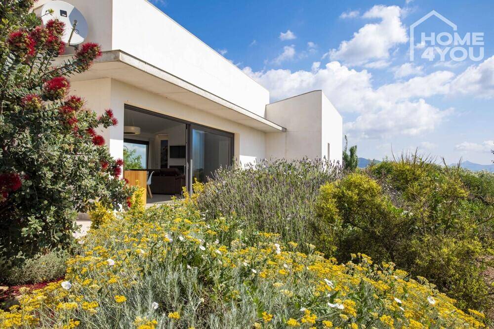 Moderne Hausfassade umgeben von blühenden Sträuchern und mediterraner Vegetation unter blauem Himmel.