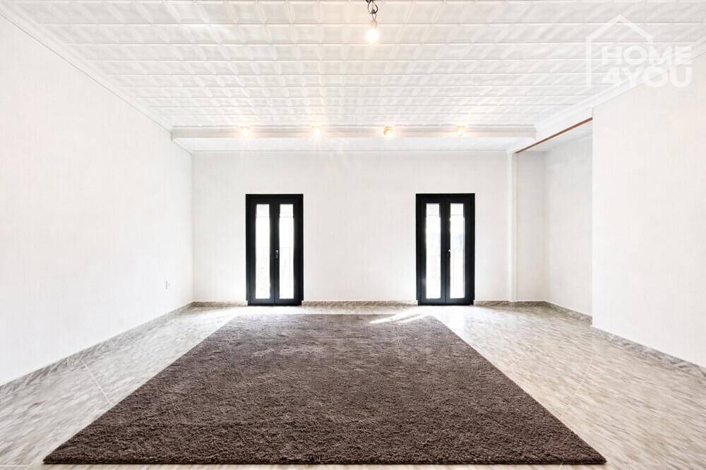 Geräumiger, heller Raum mit hohen Decken, dekorativer Deckenverkleidung und zwei dunklen Fenstertüren.