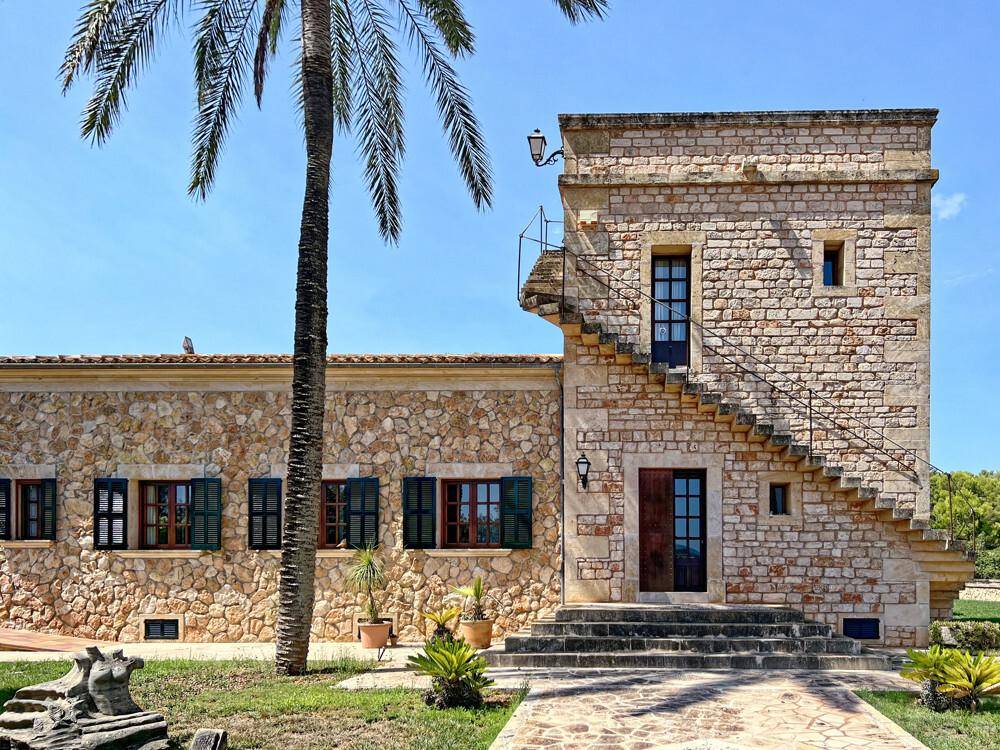 Traditionelles Steinhaus mit markantem Turm und Außentreppe, umgeben von mediterraner Vegetation.