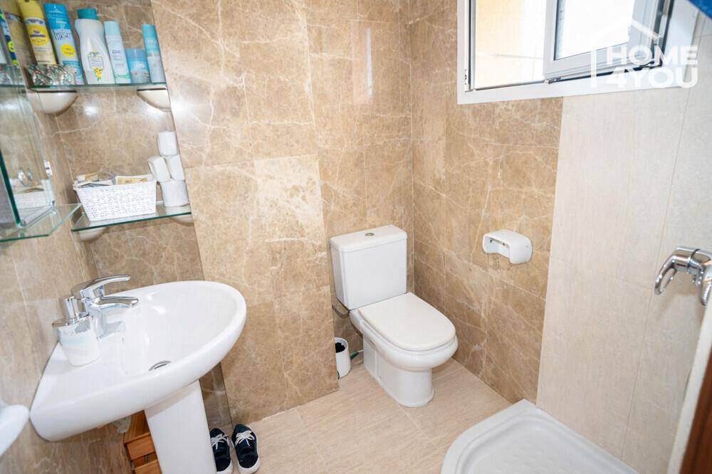Kleines WC mit beigefarbenen Fliesen, Eckwaschbecken, Toilette und Fenster.
