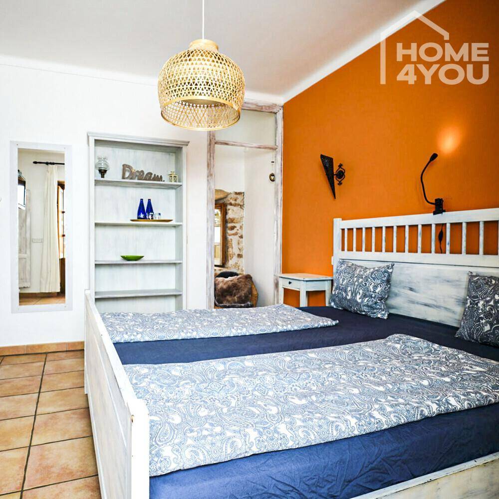 Schlafzimmer mit orangefarbener Akzentwand, weißem Bett und rustikalem Fliesenboden.