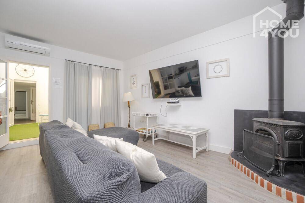 Helles Wohnzimmer mit großem Sofa, Wandfernseher und einem gemütlichen Holzofen.