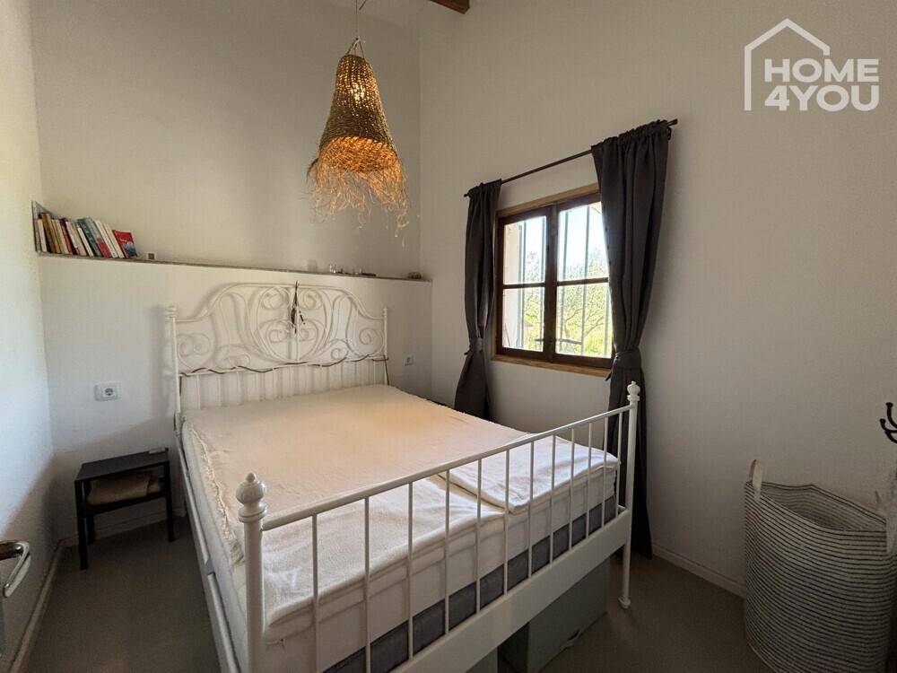 Helles Schlafzimmer mit weißem Metallbett, Holzbalken und Fenster mit Blick nach draußen.