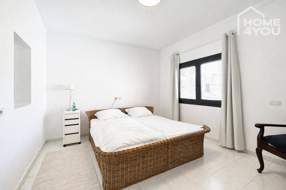 Helles Schlafzimmer mit Rattanbett, weißem Nachttisch und Fenster mit Jalousien.