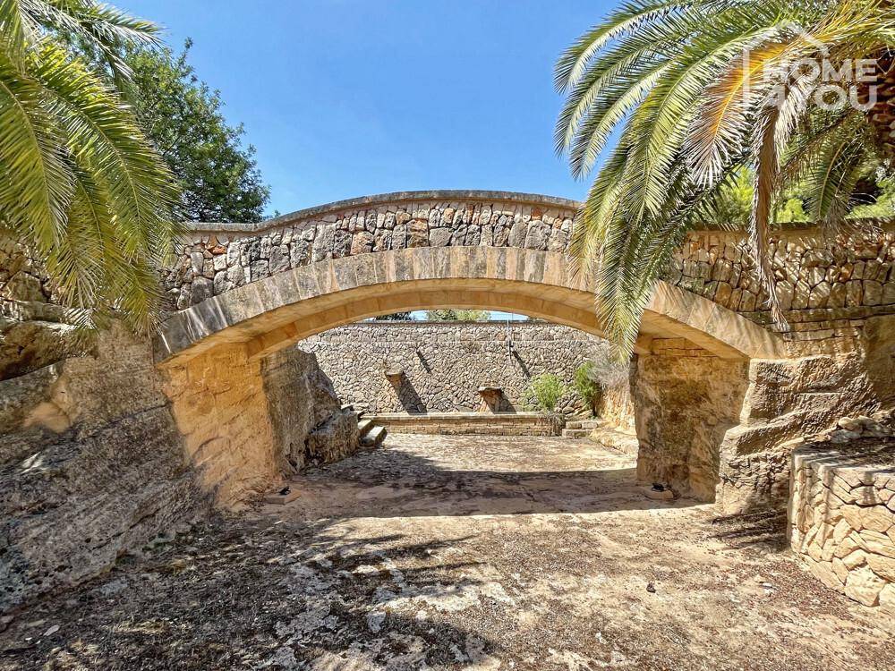 Malerische Steinbrücke im weitläufigen Gartenbereich, umgeben von mediterraner Vegetation.