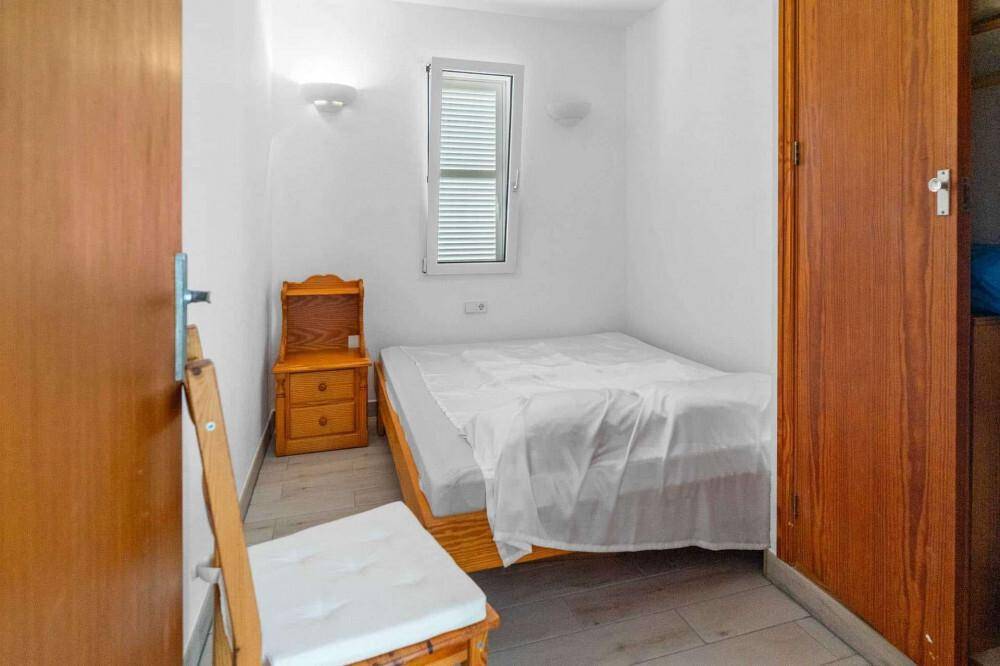 Kleines Schlafzimmer mit Einzelbett, Nachttisch und Holzmöbeln, hell beleuchtet durch ein Fenster.