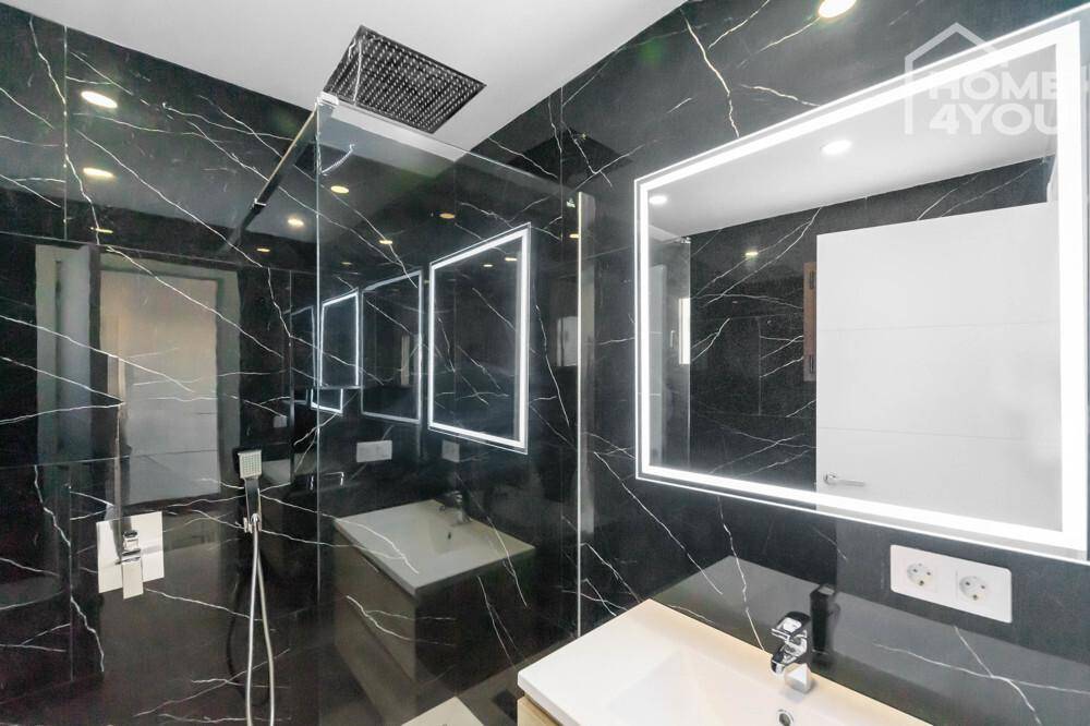 Luxuriöses Badezimmer mit schwarzen Marmorfliesen, begehbarer Dusche und beleuchtetem Spiegel.