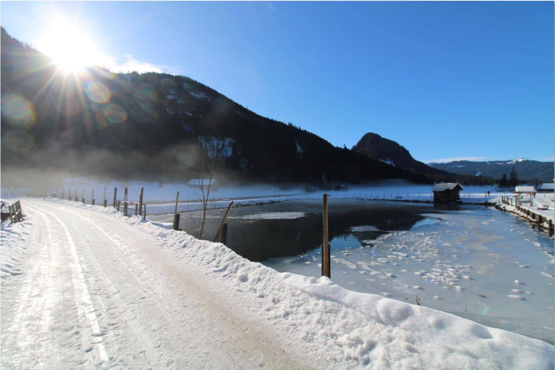 Idyllische Winterlandschaft mit gefrorenem See, verschneiter Straße und sonnigen Bergen im Hintergrund.