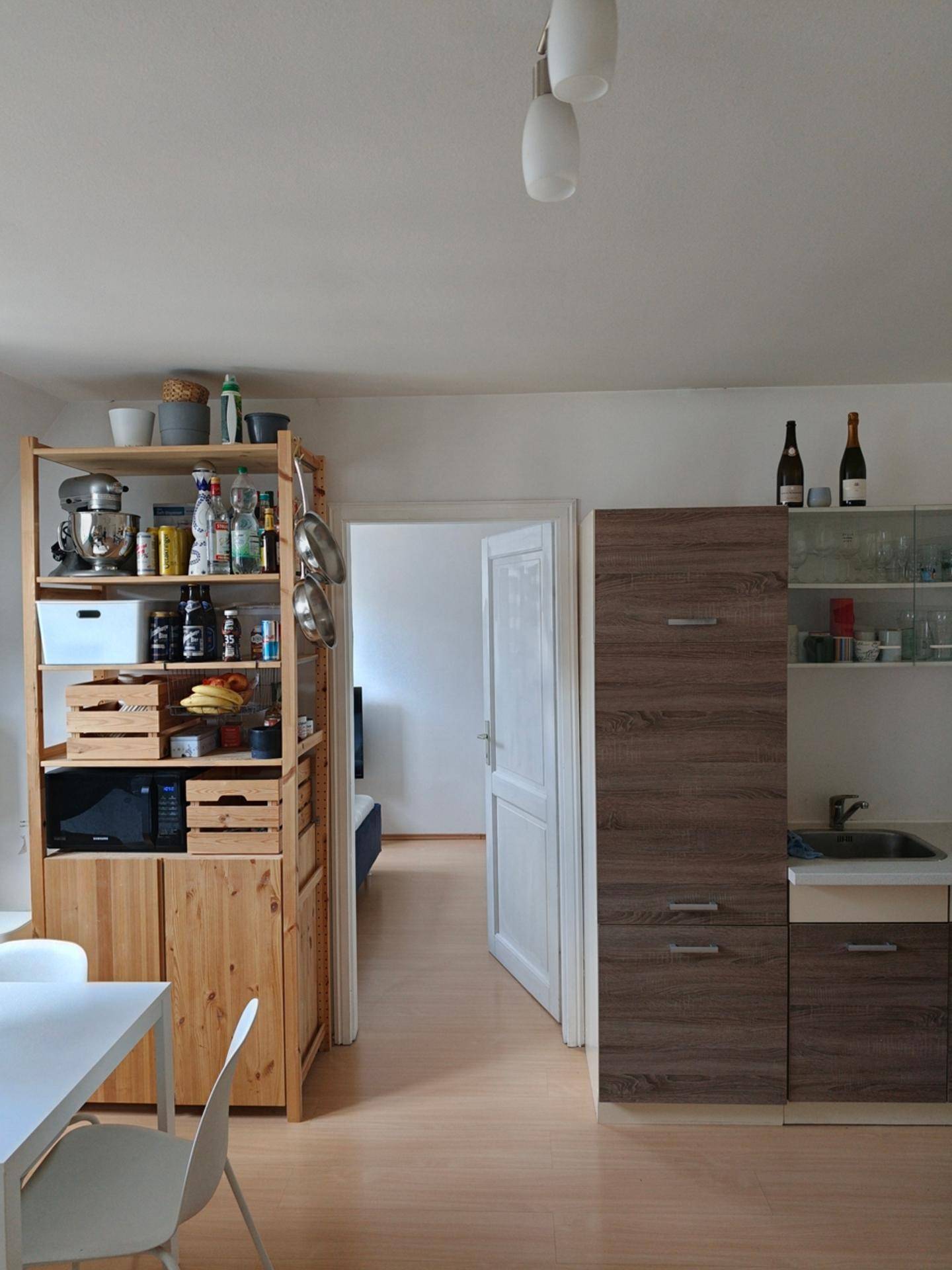 Blick von der Wohnküche in ein angrenzendes Zimmer, mit kleiner Küchenzeile und Holzregal.