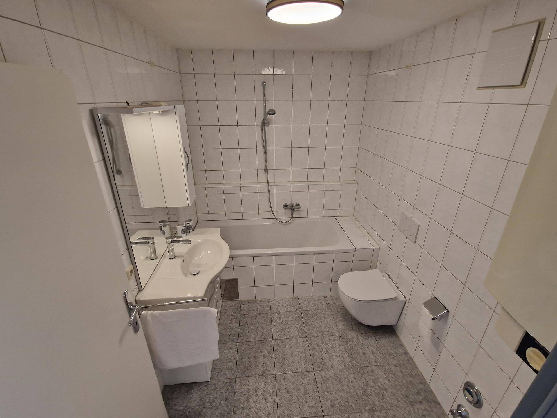 Gefliestes Badezimmer mit Badewanne, Duschfunktion, WC und Waschbecken mit Spiegelschrank.