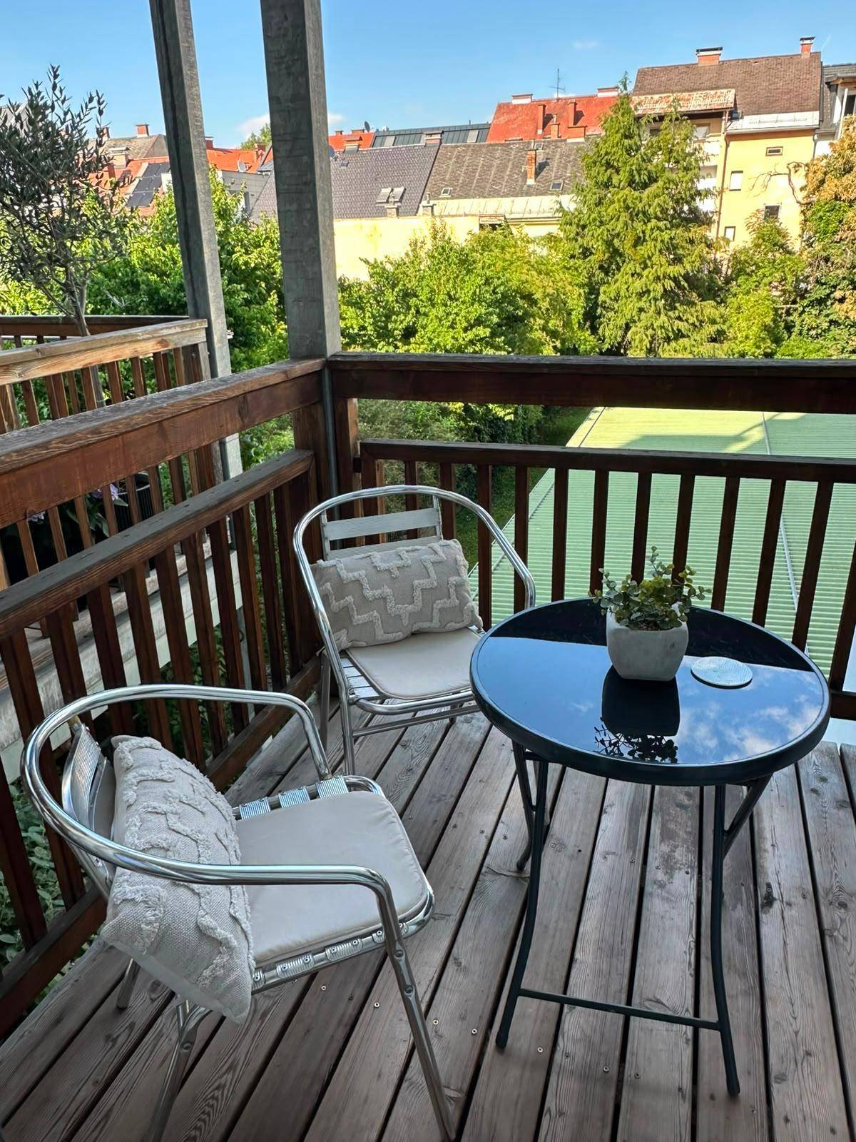 Sonniger Balkon mit Holzboden, Sitzgelegenheiten und Blick auf Bäume und Dächer.