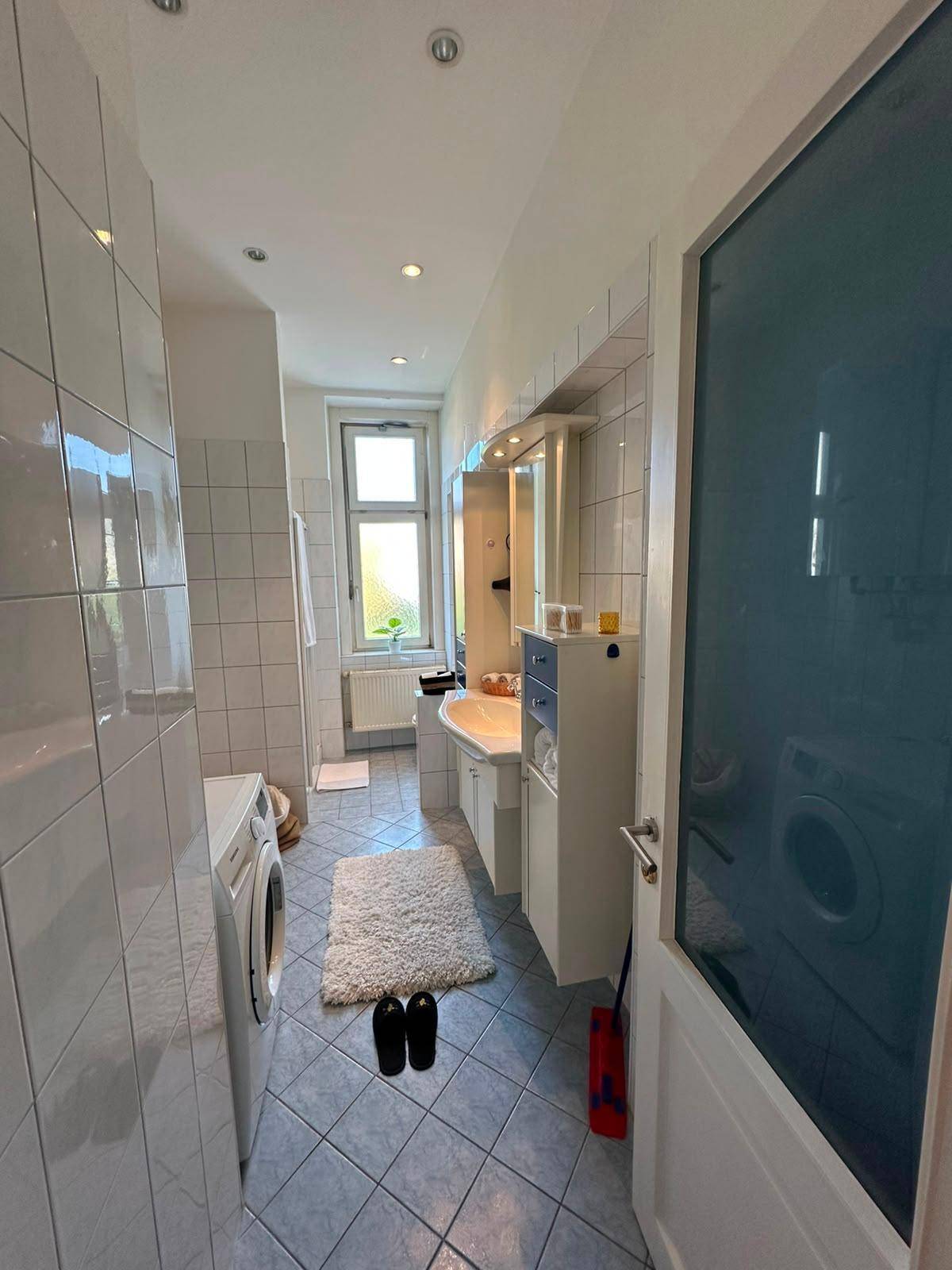 Langes Badezimmer mit Waschmaschine, WC, Waschtisch und Fenster, gefliest in Blau und Weiß.