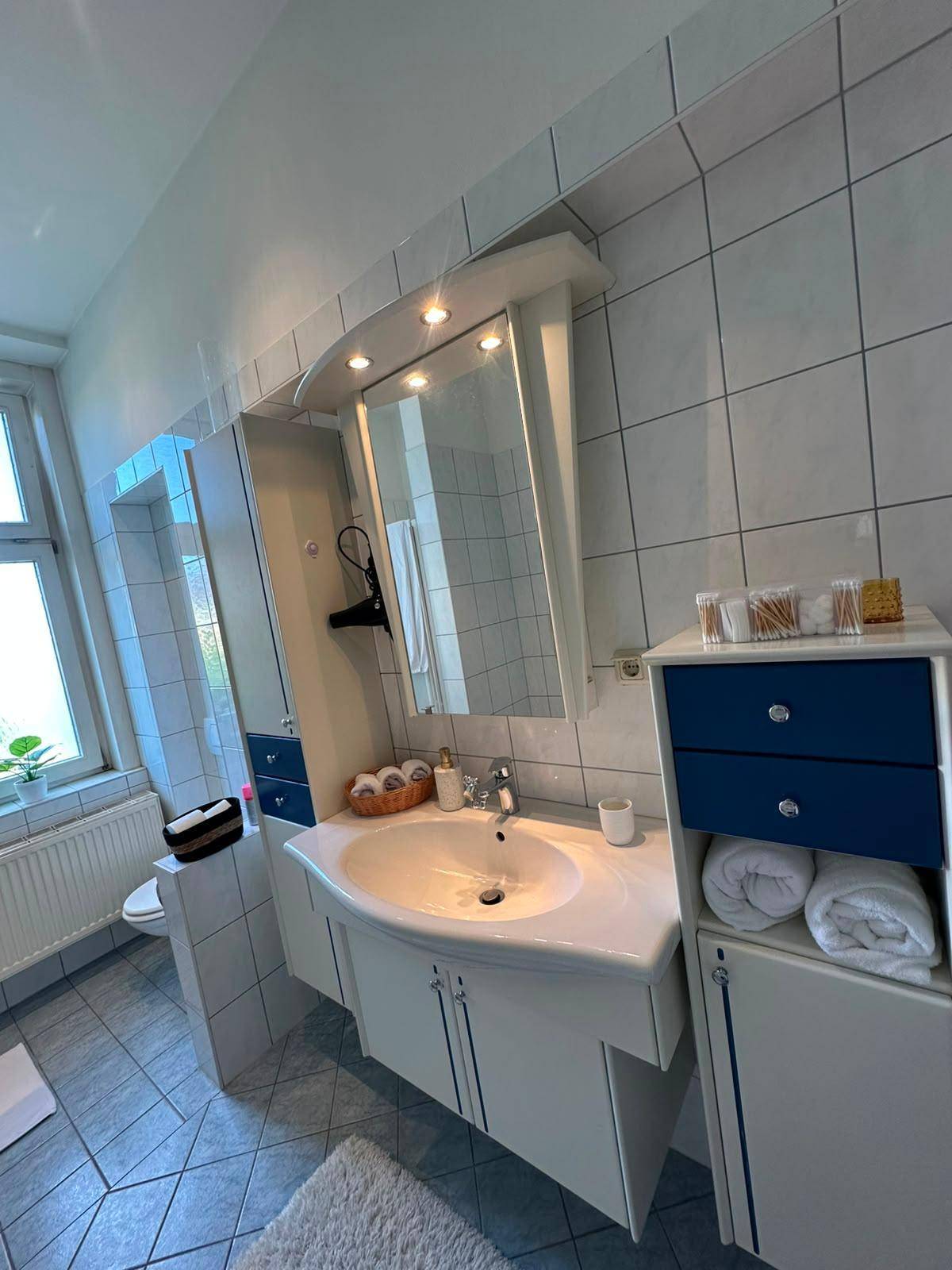 Badezimmer mit Waschbecken, Spiegelschrank, WC und Fenster, gefliest in Blau und Weiß.