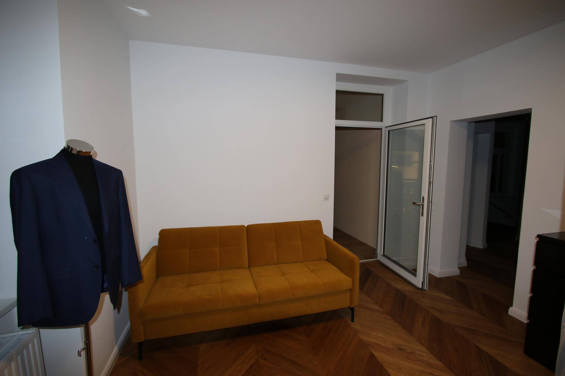 Wohnbereich mit senfgelbem Sofa, Fischgrätparkett und Zugang zu einem weiteren Raum.