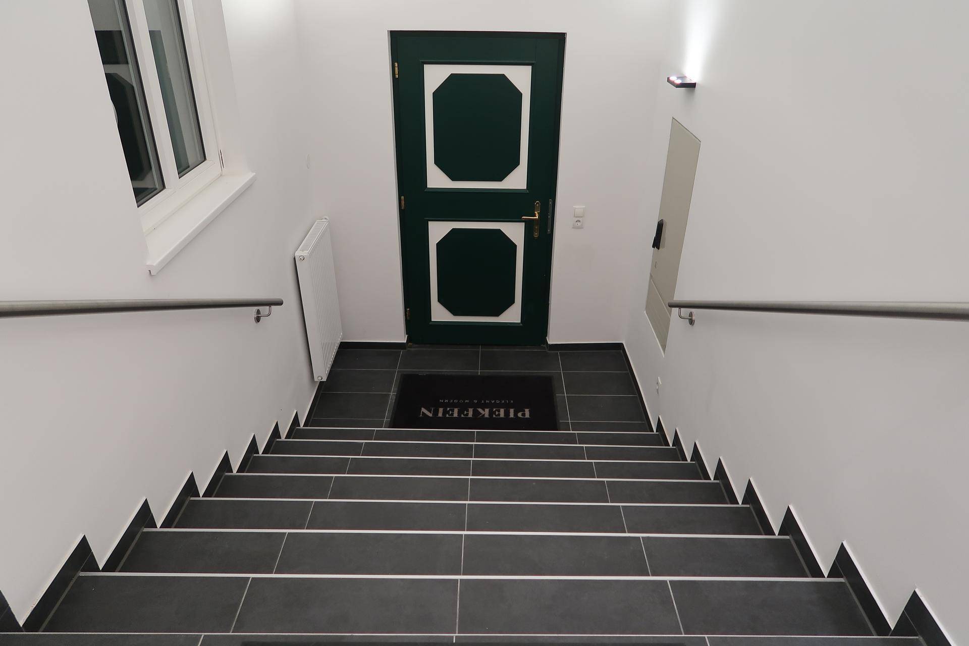 Treppenhaus mit dunklen Fliesenstufen, Metallgeländer und einer grünen Tür am oberen Ende.