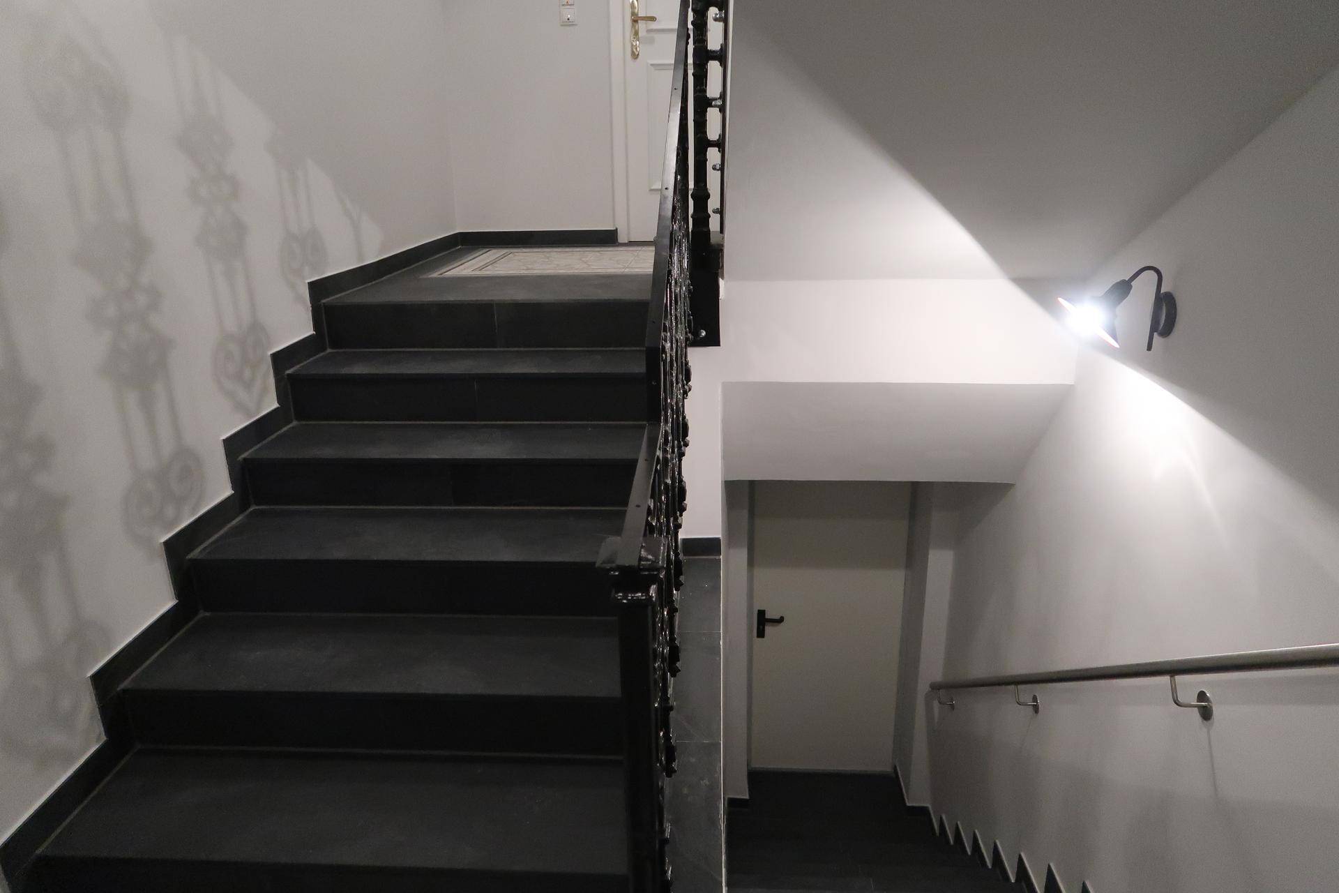 Treppenhaus mit dunklen Fliesenstufen, schmiedeeisernem Geländer und Wandbeleuchtung.