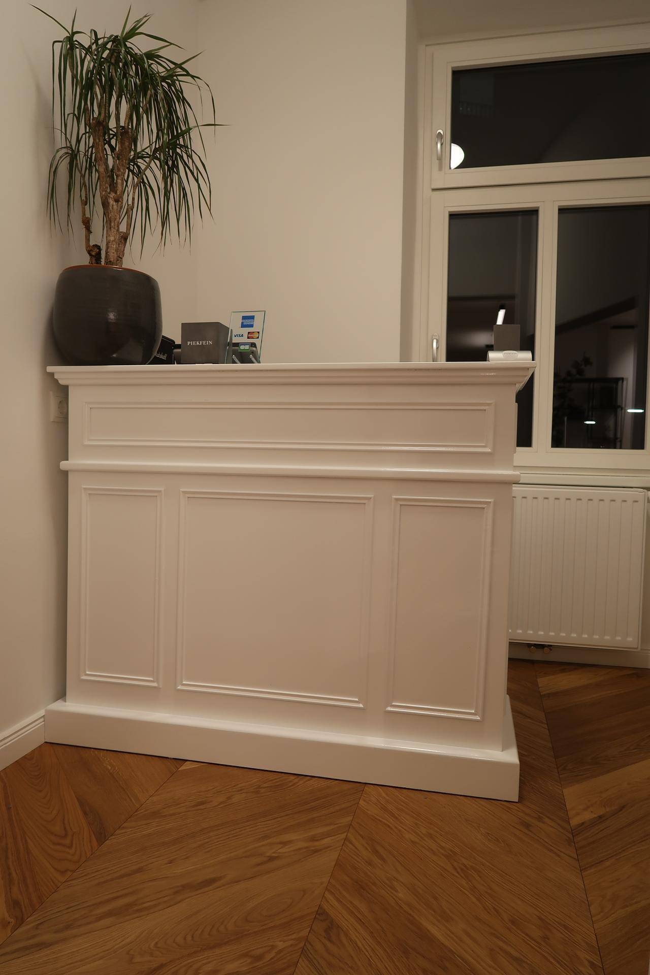 Weißer Empfangstresen mit klassischem Design, Fenster und einer Topfpflanze.
