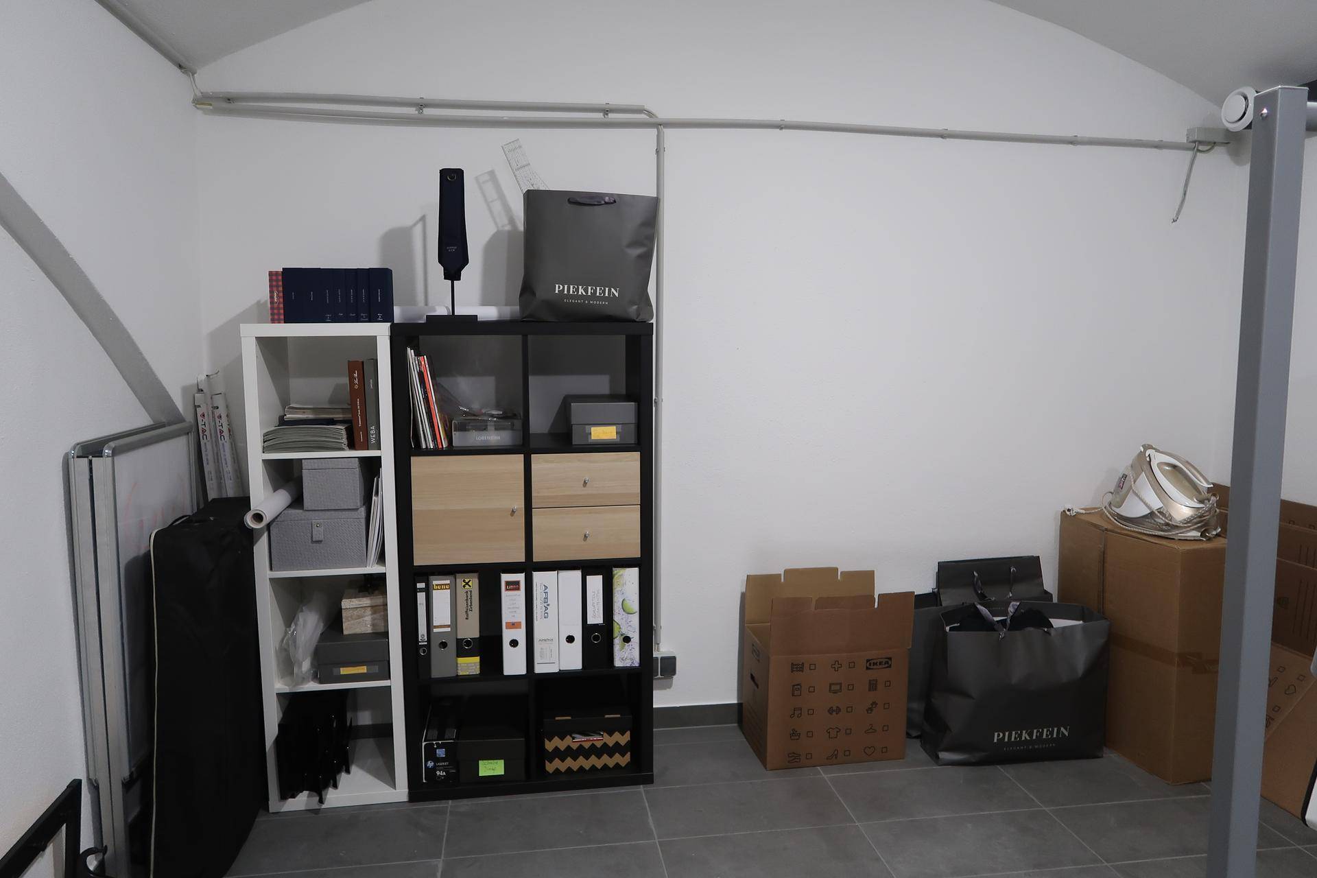 Einfacher Lagerraum mit modularen Regalen, gefüllt mit Büromaterialien und Kartons, auf grauem Fliesenboden.