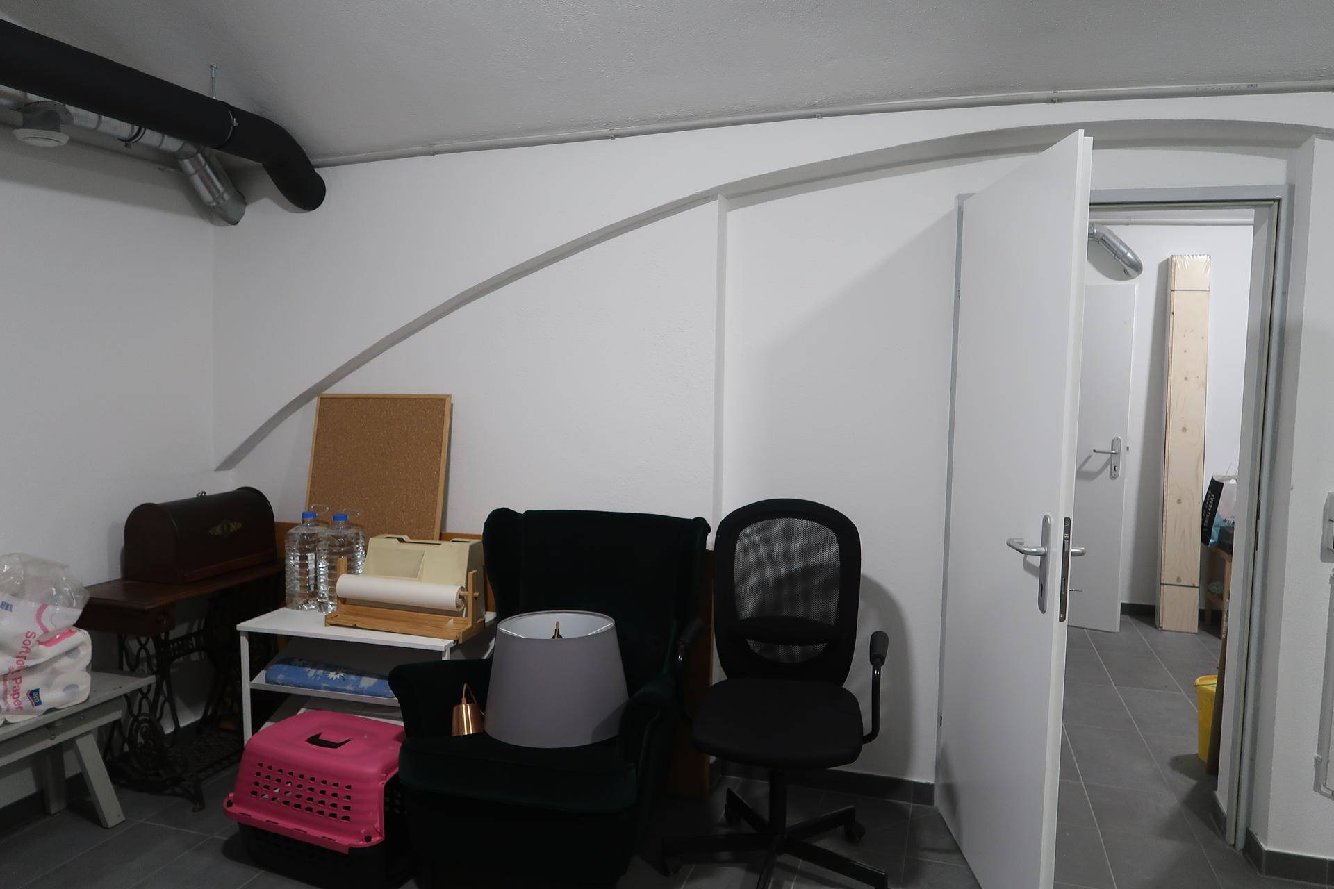 Bereich eines Mehrzweckraums mit offener Tür, Sessel, Bürostuhl und einem kleinen Tisch mit Arbeitsmaterialien.