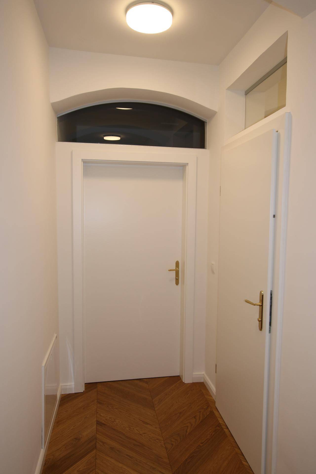 Heller Flur mit Fischgrätparkett, weißen Türen und einem Fenster über dem Türrahmen.