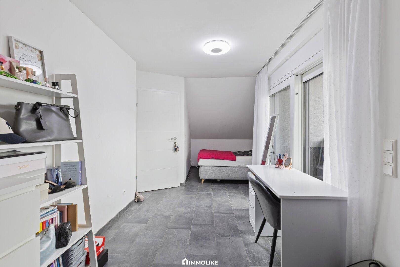 Helles Zimmer im Dachgeschoss mit Schreibtisch, Regal und Zugang zu einem kleinen Balkon.