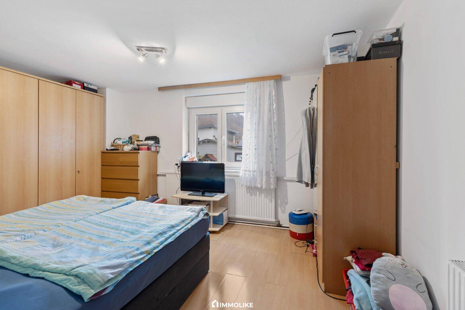Schlafzimmer mit Doppelbett, Holzschrank und Fenster, das viel Tageslicht hereinlässt.