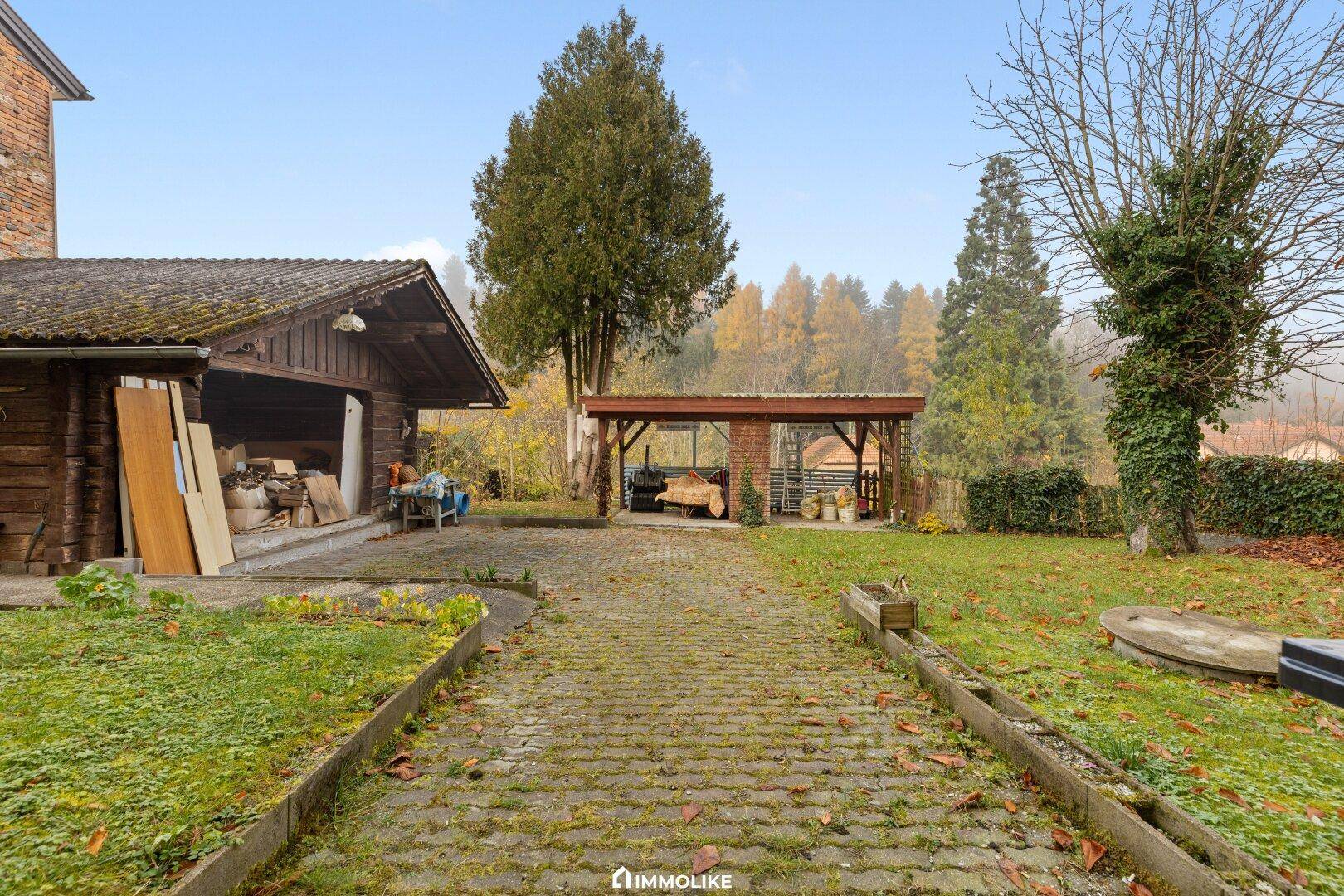 Großer Gartenbereich mit gepflasterter Zufahrt, Carport und Nebengebäuden im Grünen.