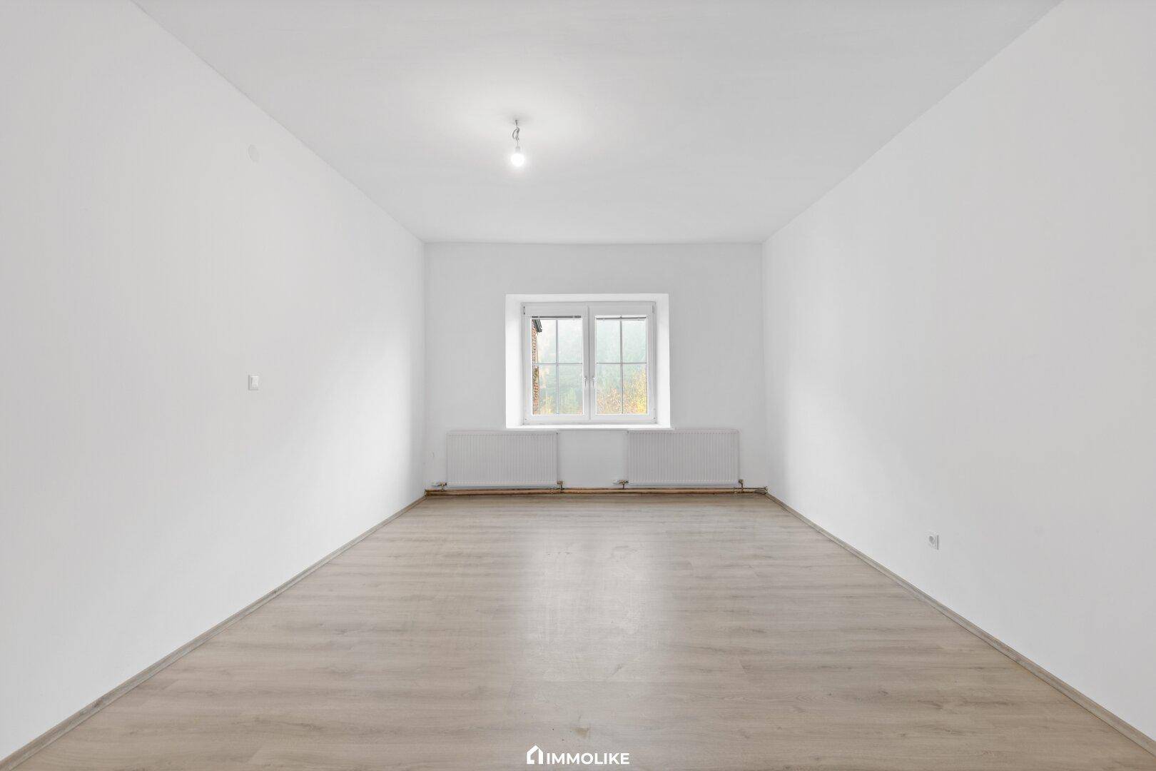 Heller, leerer Raum mit Laminatboden und einem Fenster, das viel Tageslicht hereinlässt.