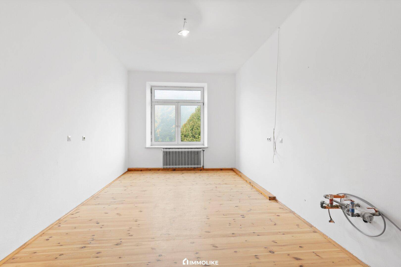 Leerer Raum mit weißen Wänden, Holzfußboden und einem Fenster mit Heizkörper darunter. Sichtbare Installationen an der Wand.