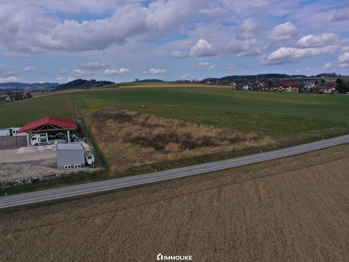 Panoramische Luftaufnahme des Gebäudes mit rotem Dach, angrenzenden Feldern und einer Landstraße unter bewölktem Himmel.