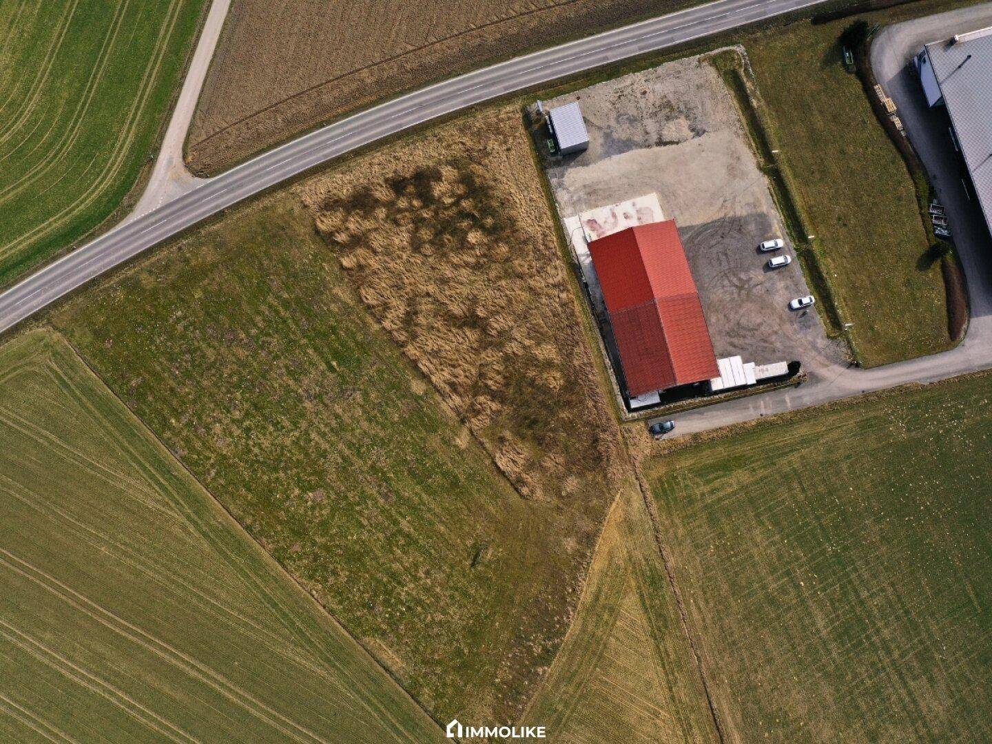 Gedrehte Luftaufnahme eines Gebäudes mit rotem Dach, umgeben von Ackerland und einer Straße, die das Grundstück begrenzt.