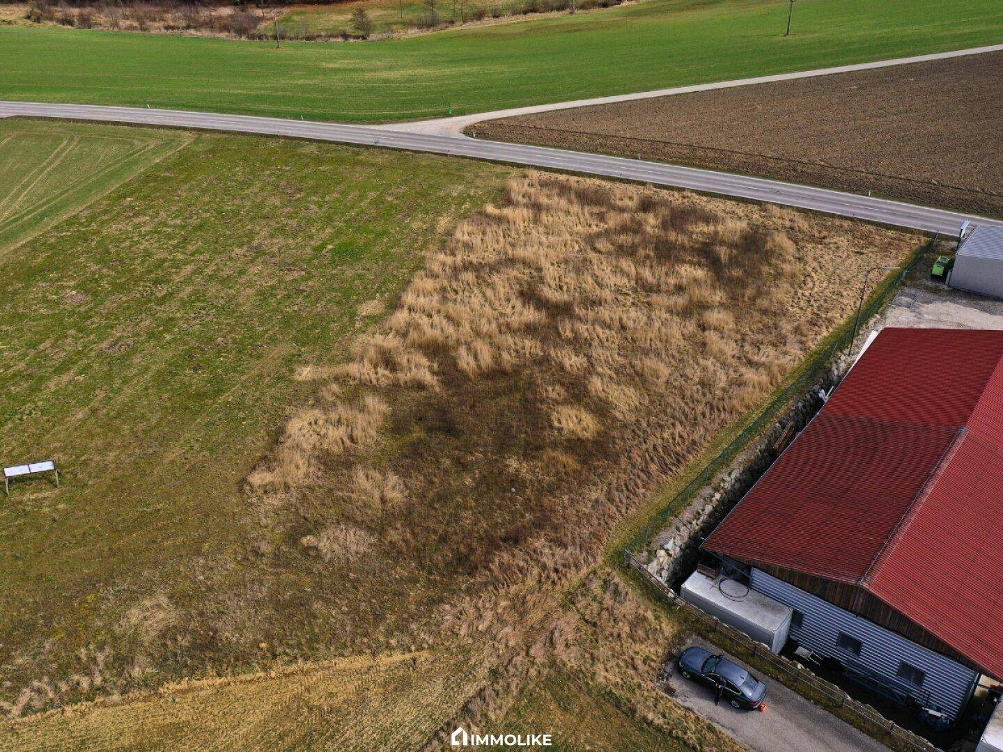 Nähere Luftaufnahme eines unbebauten Grundstücks neben einem Gebäude mit rotem Dach, begrenzt durch eine Landstraße.