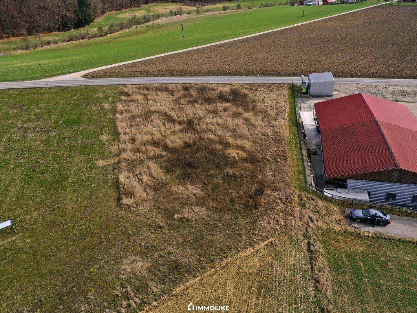 Luftaufnahme des unbebauten Grundstücks neben einem Gebäude mit rotem Dach und angrenzender Straße in ländlicher Umgebung.