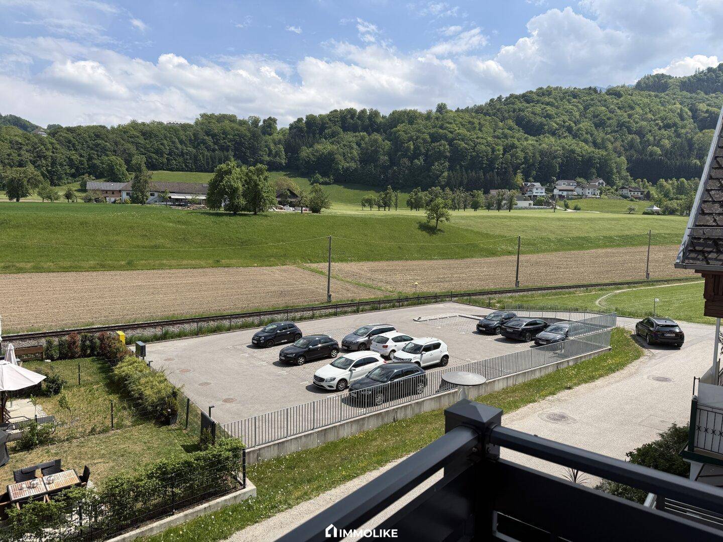 Großer Parkplatz mit mehreren Fahrzeugen und weitem Blick auf grüne Hügel und Felder in der Umgebung.