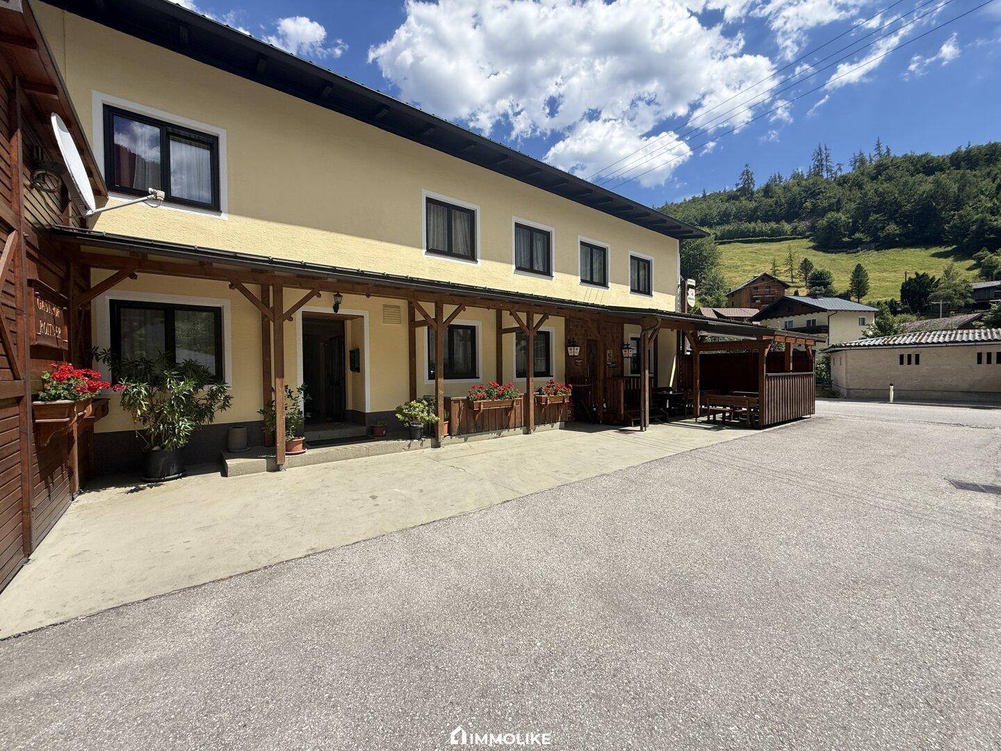 Gasthaus mit gelber Fassade, Holzbalken und überdachtem Außenbereich vor einer Bergkulisse.
