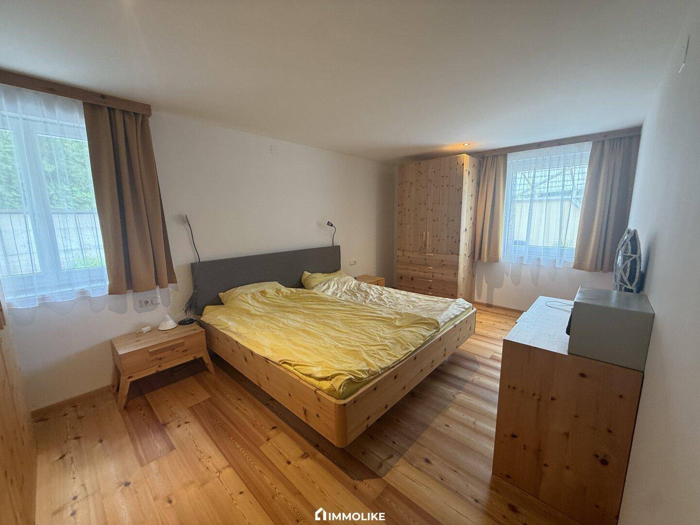 Helles Schlafzimmer mit Holzboden, Doppelbett und zwei Fenstern, die viel Tageslicht hereinlassen.