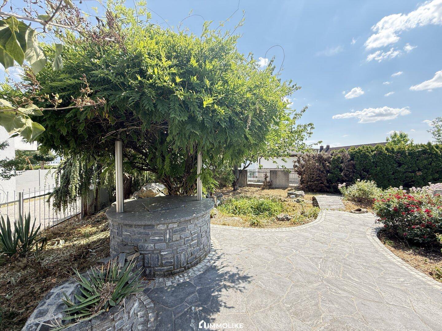 Dekorativer Brunnen im Gartenbereich, umgeben von üppigem Grün und einem gepflasterten Weg.