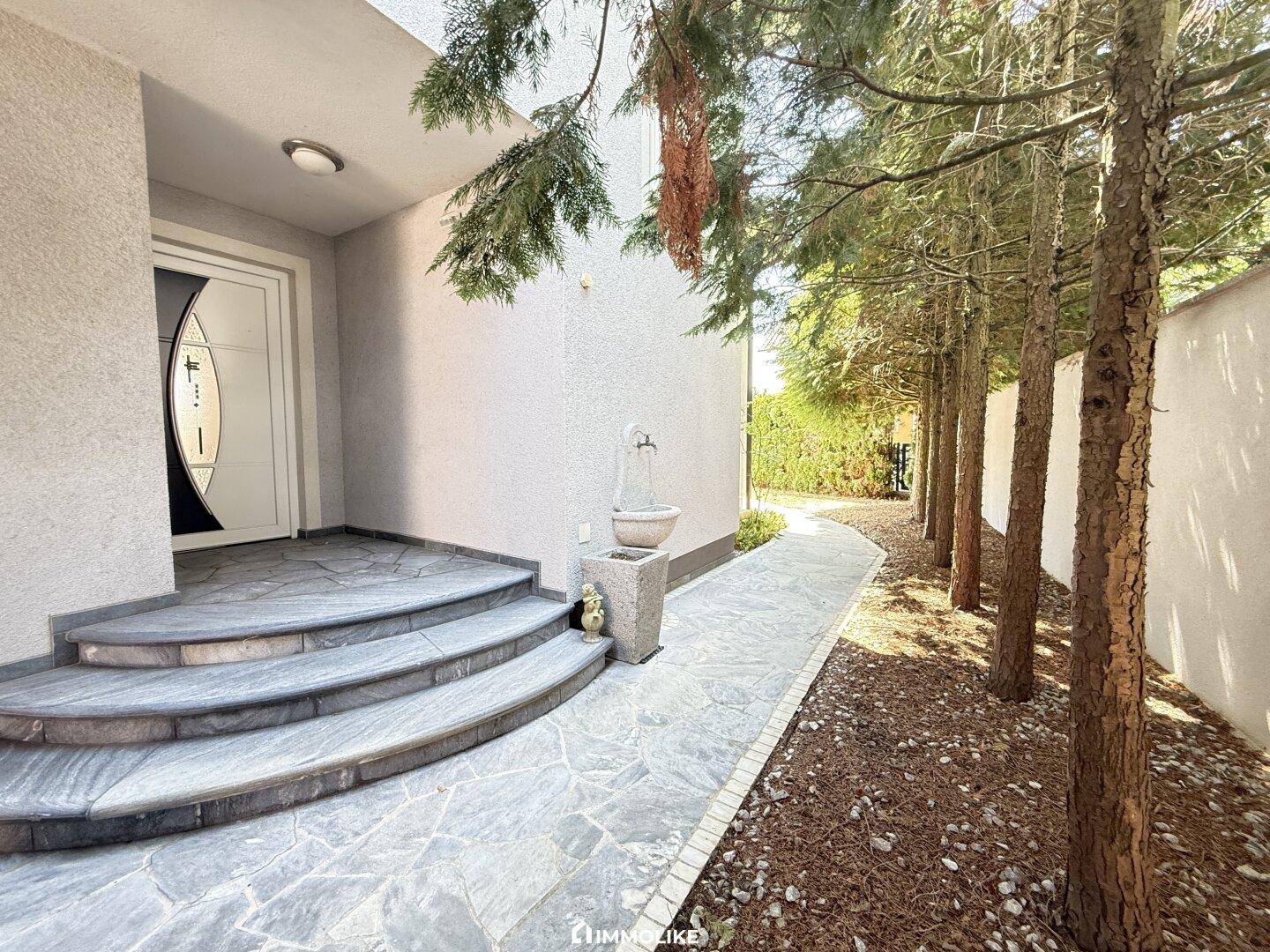 Eingangsbereich des Hauses mit eleganten Steinstufen und einem gepflasterten Weg, gesäumt von Bäumen.