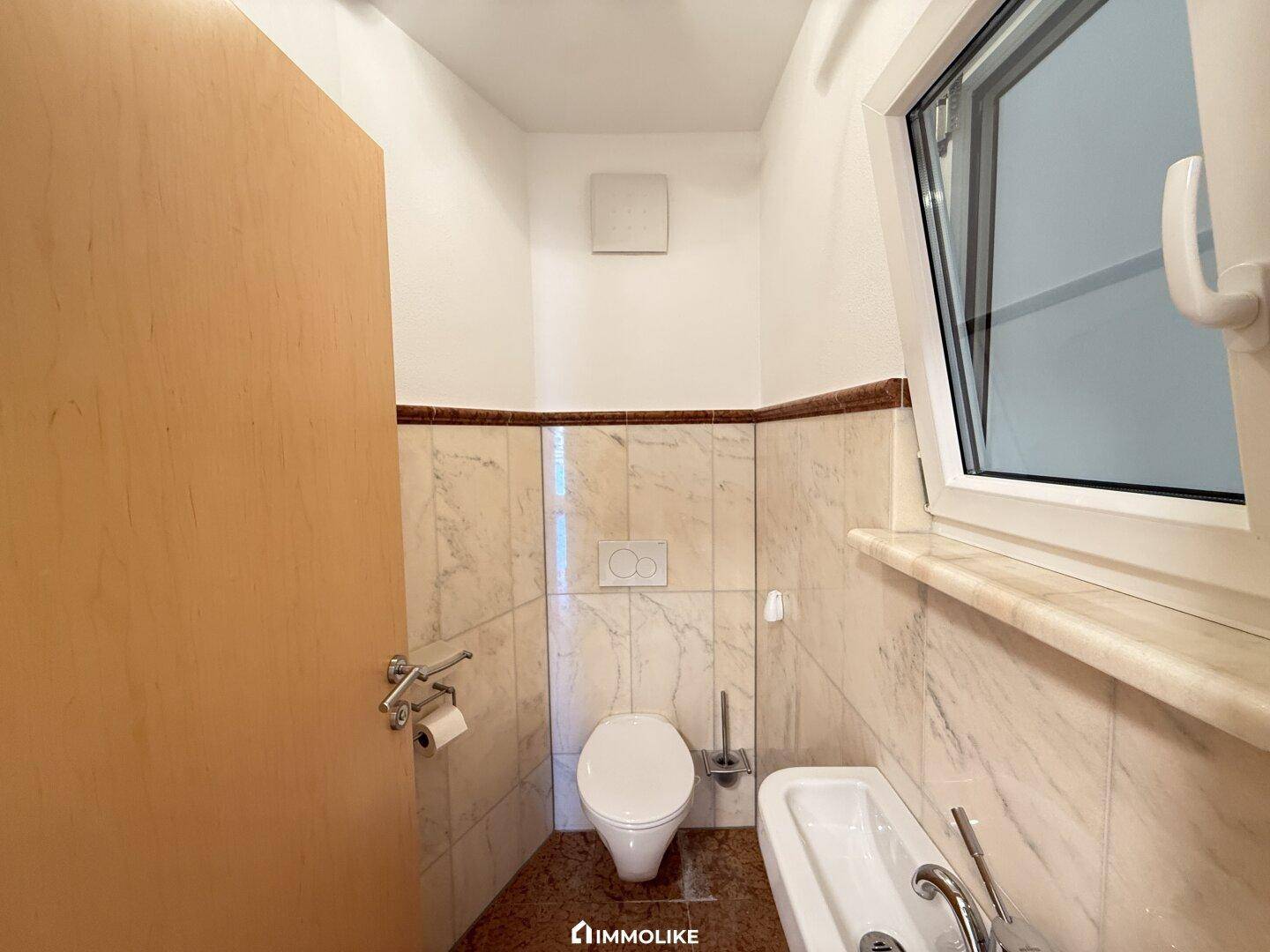 Modernes Gäste-WC mit Wand-WC, kleinem Waschbecken und Fenster für natürliche Belüftung.