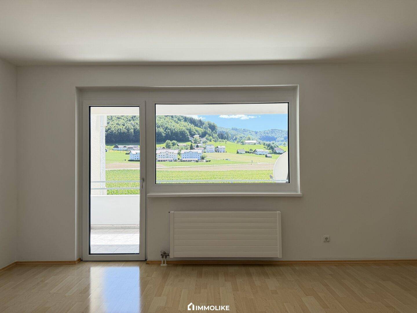 Helles Zimmer mit Parkettboden, großem Fenster und Zugang zu einem Balkon mit Landschaftsblick.