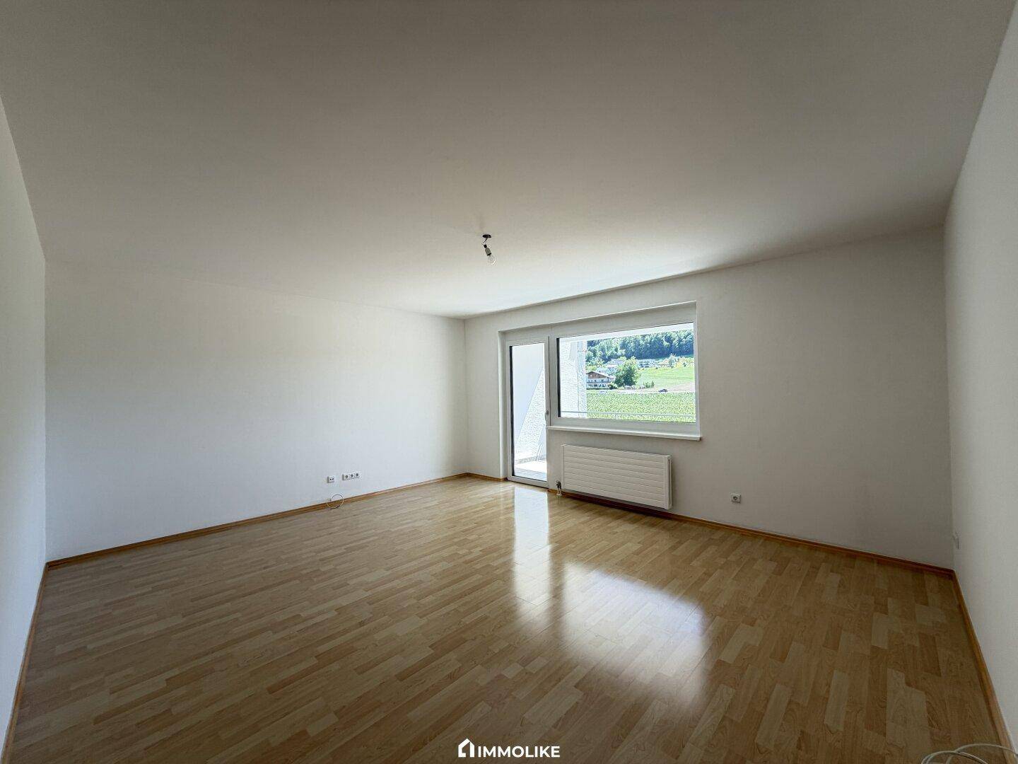 Geräumiges, leeres Zimmer mit hellem Parkettboden und viel Tageslicht durch das große Fenster.