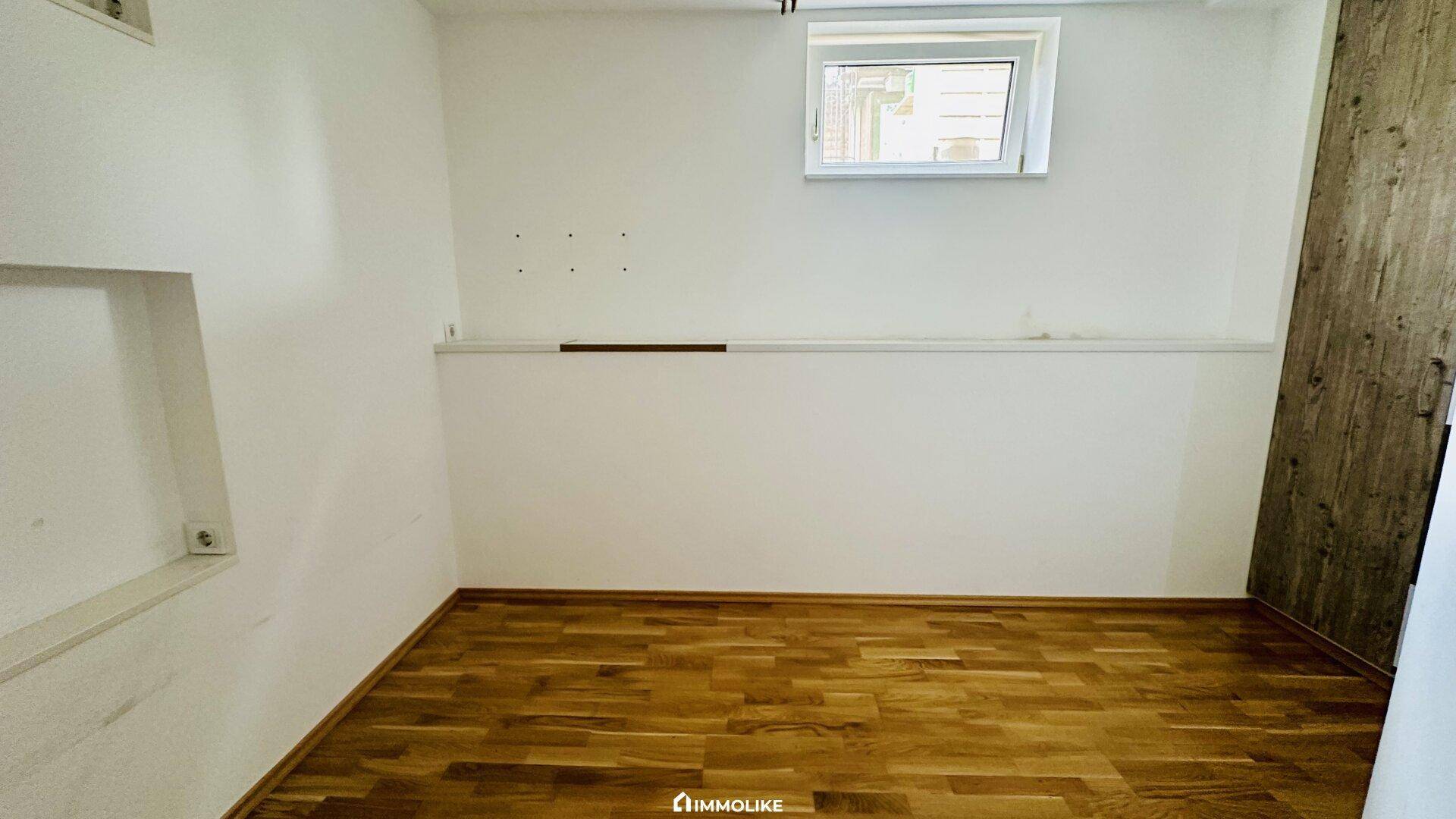 Leerer Raum mit hellem Holzboden, weißen Wänden und einem Fenster für natürliches Licht.